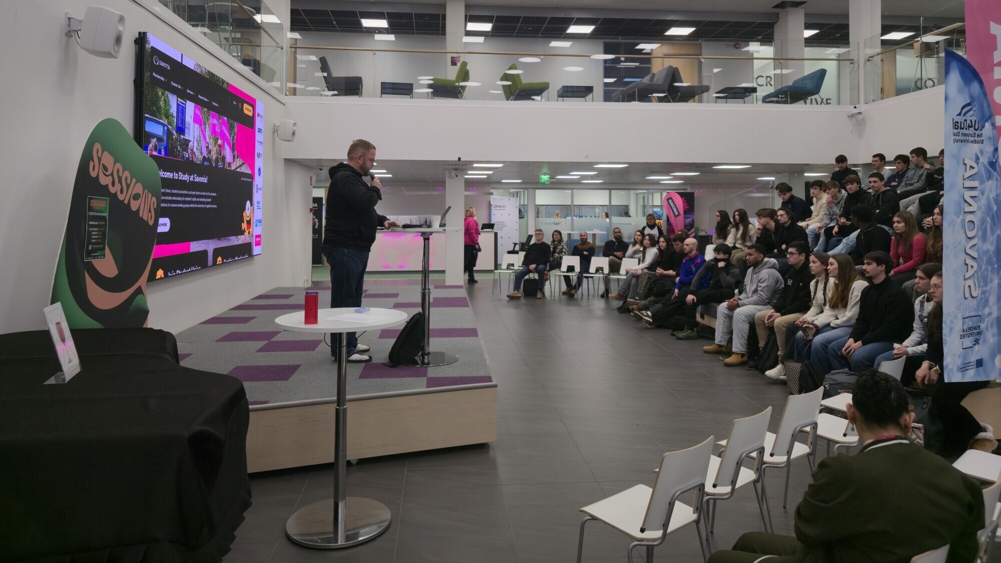 Puhuja puhuu korokkeella modernissa, tilavassa tilassa, jossa yleisö istuu valkoisilla tuoleilla. Suurella näytöllä näkyy värikästä grafiikkaa, ja banderollit koristavat aluetta. Jotkut ihmiset katsovat tilannetta ylemmältä tasolta.