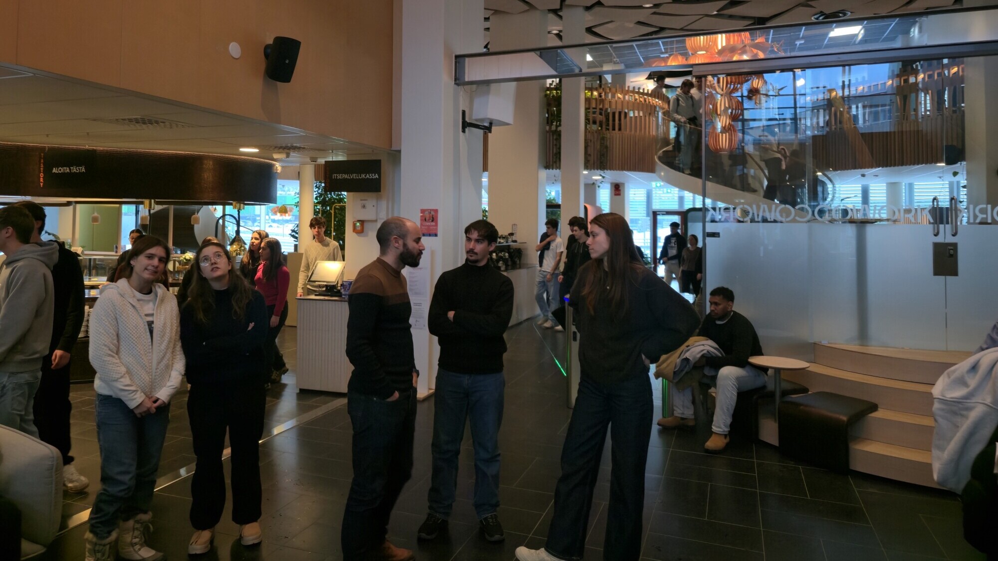 Joukko ihmisiä seisoo ja keskustelee modernissa, kirkkaasti valaistussa kahvilassa, jossa on korkeat katot, geometriset kattopaneelit ja lasiseinät. Muut ihmiset istuvat ja rentoutuvat taustalla kierreportaiden lähellä.