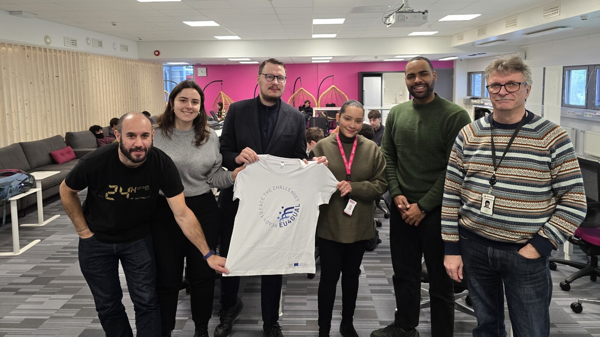 Kuusi ihmistä seisoo sisätiloissa toimistossa ja hymyilee kameralle. Kahdella heistä on valkoinen t-paita, jossa on sininen logo ja teksti Erasmus+ Challenge. Muut ihmiset työskentelevät taustalla työpöydissä.
