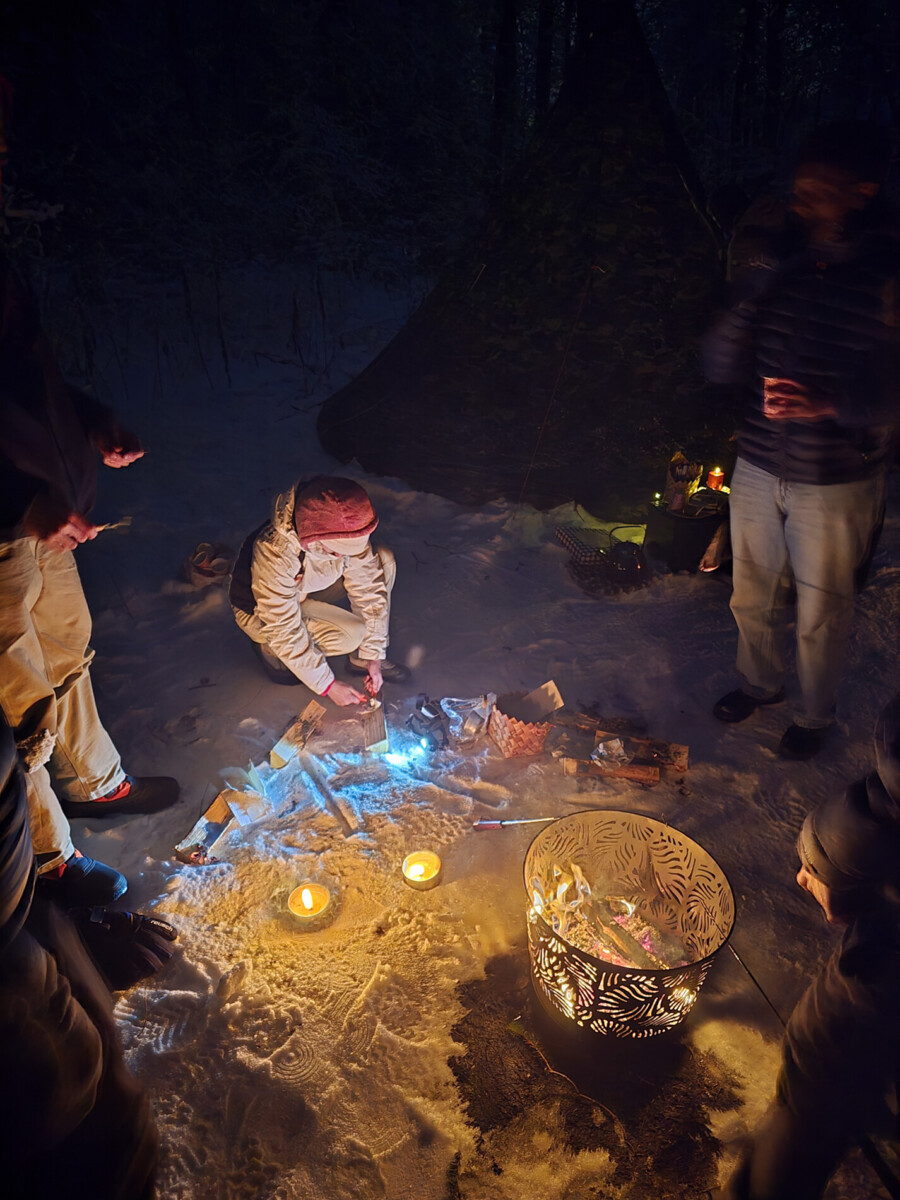 Henkilö polvistuu lumisella maalla ja hoitaa pientä nuotiota, jota ympäröivät sytytetyt kynttilät, ja useita muita seisoo lähellä. Hämärästi valaistussa yöllisessä taustassa näkyy teltta ja puita.