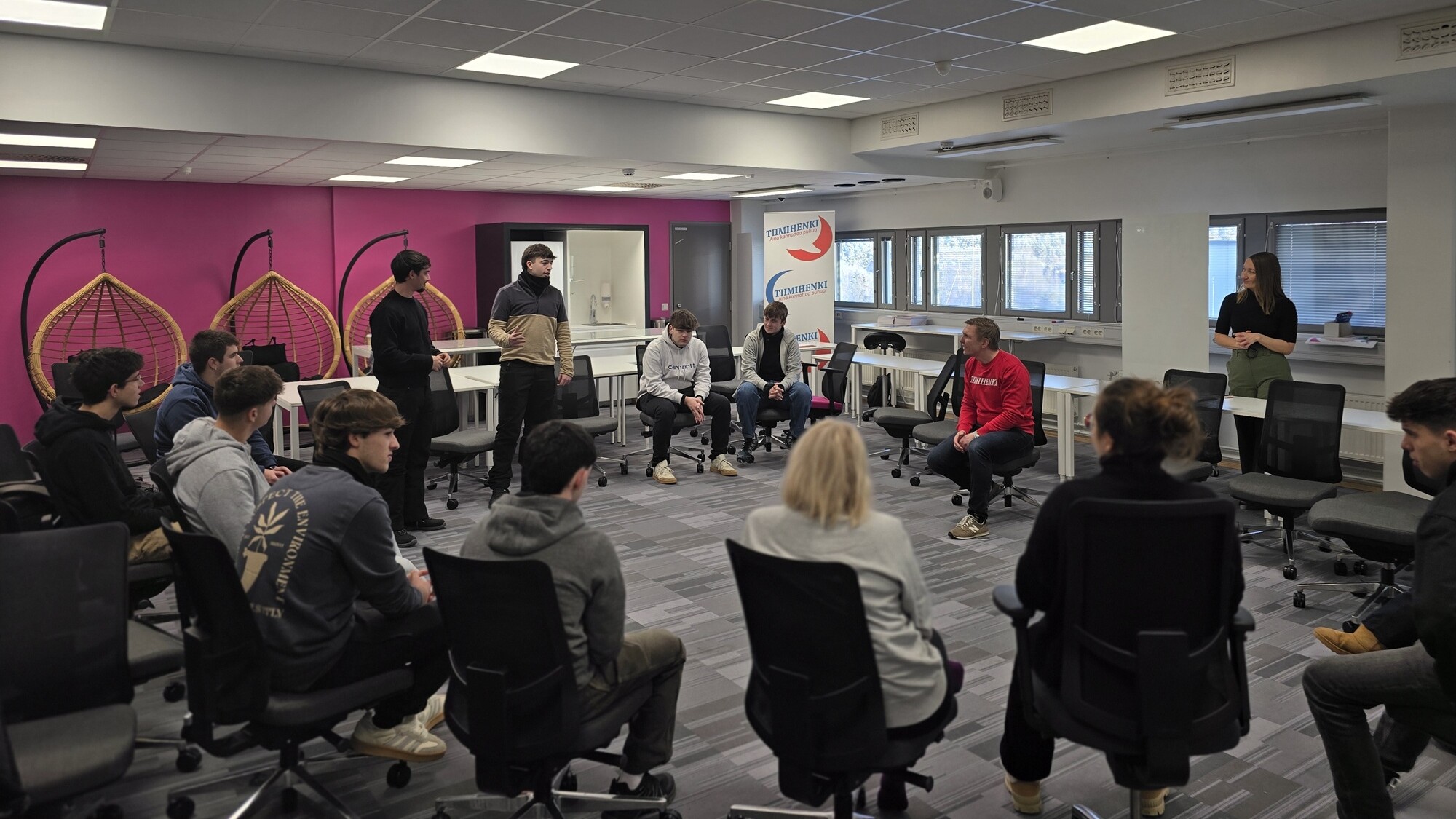 Joukko ihmisiä istuu tuolien muodostamassa piirissä modernissa, kirkkaasti valaistussa huoneessa, jonka seinät ovat vaaleanpunaiset ja valkoiset. Jotkut osallistujat puhuvat tai kuuntelevat, kun taas yksi henkilö seisoo taulun vieressä.