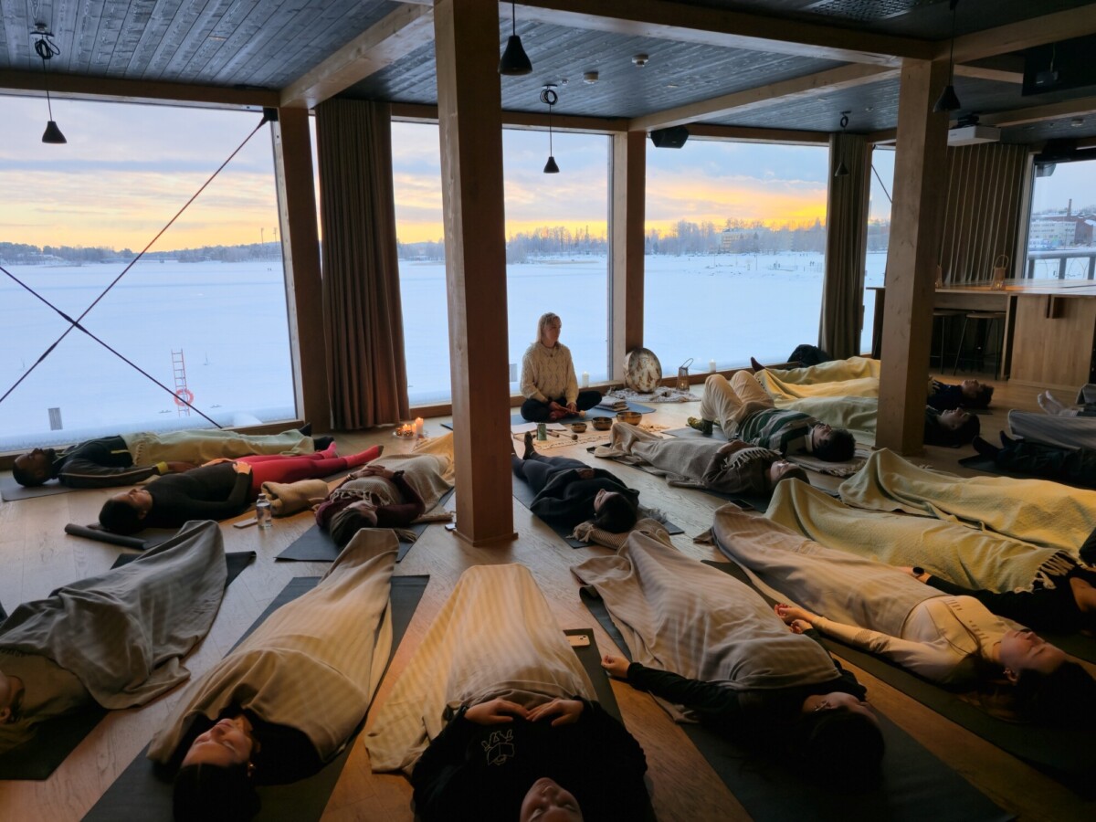 Joukko ihmisiä makaa lattialla peittojen alla joogastudiossa, jossa on suuret ikkunat, kun ohjaaja istuu keskellä. Ulkona lumi peittää maiseman, ja aurinko laskee ja heittää lämpimän hehkun sisälle.