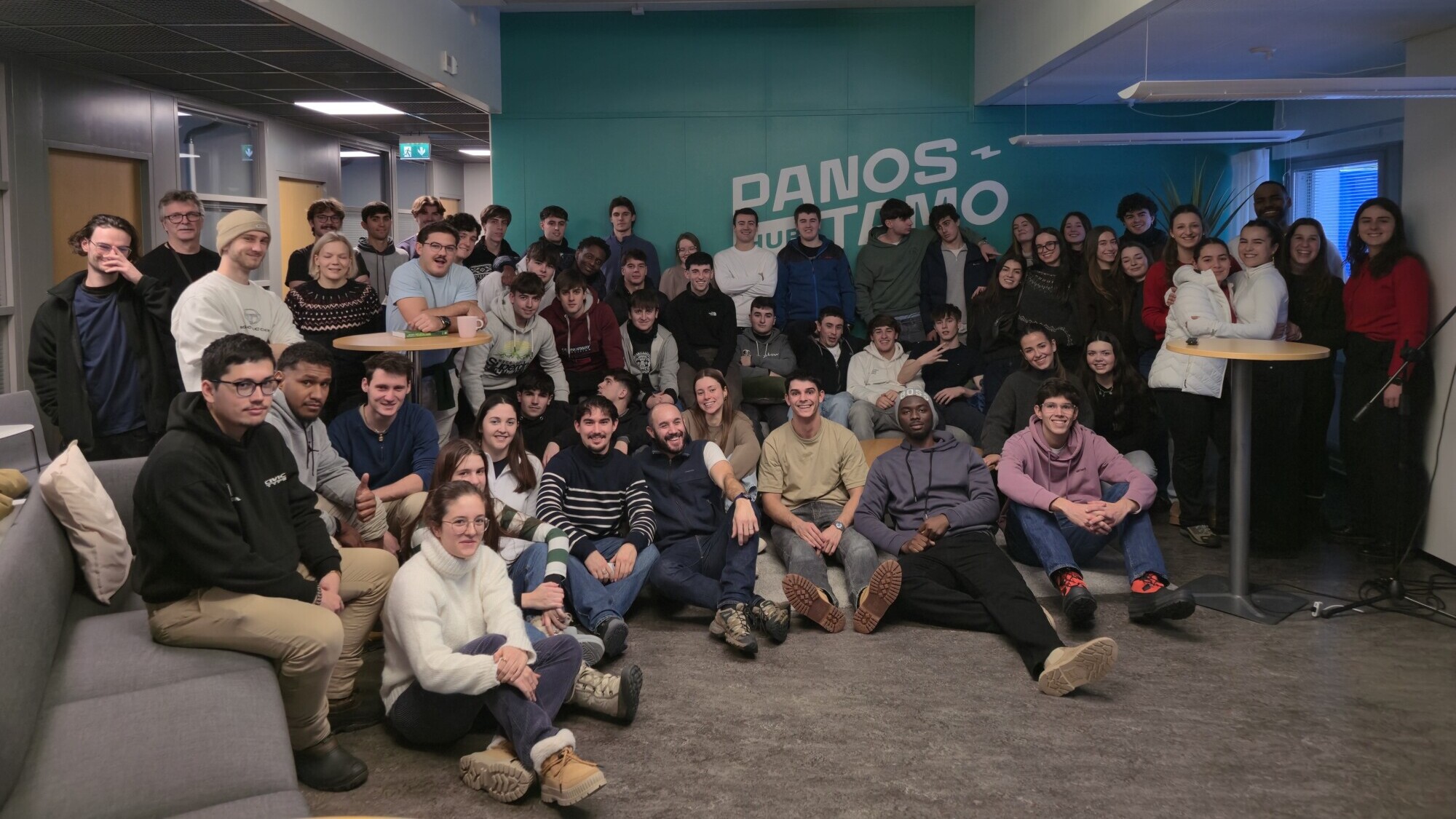 Suuri joukko ihmisiä on kokoontunut toimistotilaan ja hymyilee kameralle. He istuvat ja seisovat vaaleanpunaisen seinän edessä, jossa on osittain näkyvissä teksti DANOS CULTEMOS.