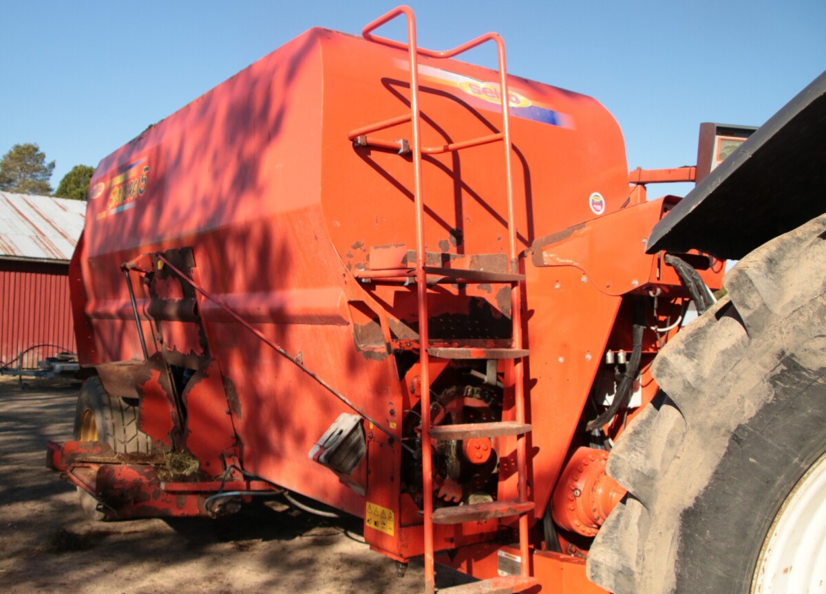 Traktoriin on kiinnitetty suuri punainen maatalouskone, todennäköisesti rehusekoitin. Metalliportaat johtavat sivulle, ja koneen alla näkyy osittain henkilö. Taustalla on vaja ja puita.