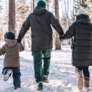 Kolme ihmistä, kaksi aikuista ja lapsi, kävelevät käsi kädessä lumisessa metsässä. Heillä on yllään talvitakit, pipot ja saappaat, ja heidät nähdään takaapäin, kun he kävelevät korkeiden puiden seassa.