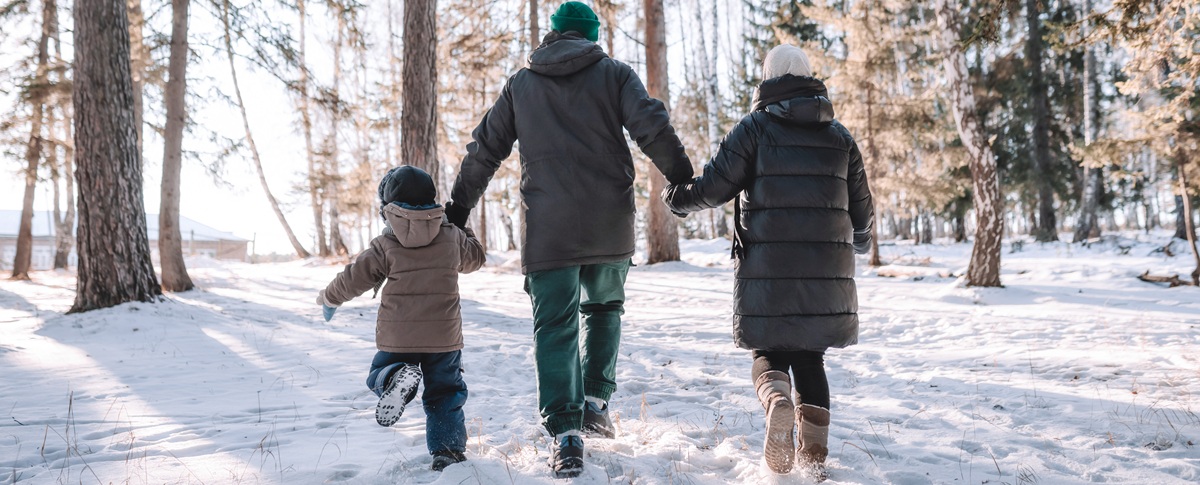 Kolme ihmistä, kaksi aikuista ja lapsi, kävelevät käsi kädessä lumisessa metsässä. Heillä on yllään talvitakit, pipot ja saappaat, ja heidät nähdään takaapäin, kun he kävelevät korkeiden puiden seassa.