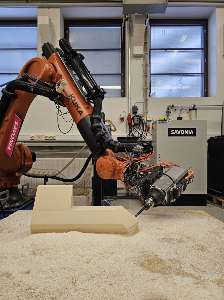 Teollinen KUKA-robottikäsivarsi veistää materiaalilohkoa, jolloin työpajan lattialle syntyy lastuja. Robotti on merkitty nimellä "Savonia", ja se työskentelee lähellä ikkunaseinää, jossa on näkyvissä laitteita ja pistorasioita.