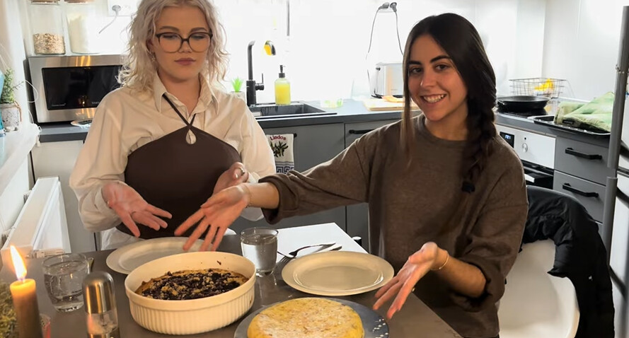 Kaksi naista istuu keittiön pöydän ääressä hymyillen ja viittoen kahta edessään olevaa ruokalajia - leivottua pataa ja pyöreää litteää leipää. Keittiö on kirkkaasti valaistu, ja taustalla on erilaisia astioita ja esineitä.