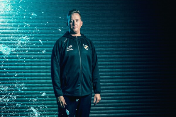 Tummiin urheilupukuihin pukeutunut henkilö seisoo kameraan päin, ja vasemmalla puolella on lasinsirpale-efekti ja tiilenpunainen valaistus ja oikealla puolella tumma, harjakuvioinen tausta.