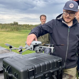 Silmälasipäinen ja lippalakkipäinen mies valmistautuu laukaisemaan dronea suuresta mustasta kotelosta ulkona, kun toinen mies seisoo taustalla ruohokentällä pilvisen taivaan alla.