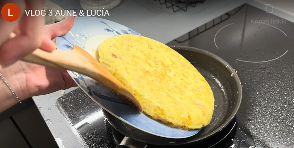 Henkilö liu'uttaa lastalla pyöreän, kullanvärisen espanjalaisen munakkaan paistinpannulta sinivalkoiselle lautaselle liedellä.