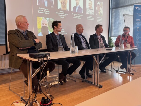 Viisi liikeasuun pukeutunutta miestä istuu paneelipöydän ääressä mikrofonien ja vesipullojen kanssa keskustelemassa. Heidän takanaan on valkokangas, jolla näkyy nimiä, titteleitä ja valokuvia.