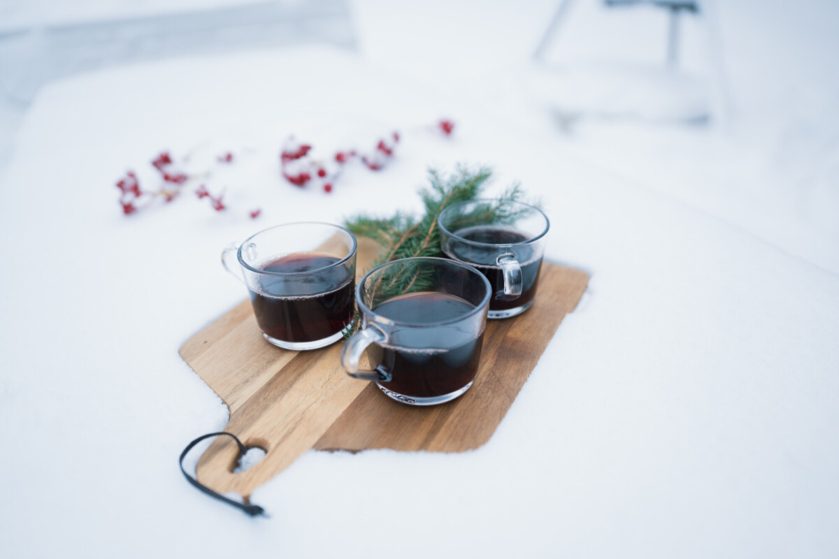 Kolme tummalla juomalla täytettyä lasimukia istuu puulautalla lumessa, ja niiden välissä on vihreä männyn oksa. Punaisia marjoja on ripoteltu sumeaan taustaan.