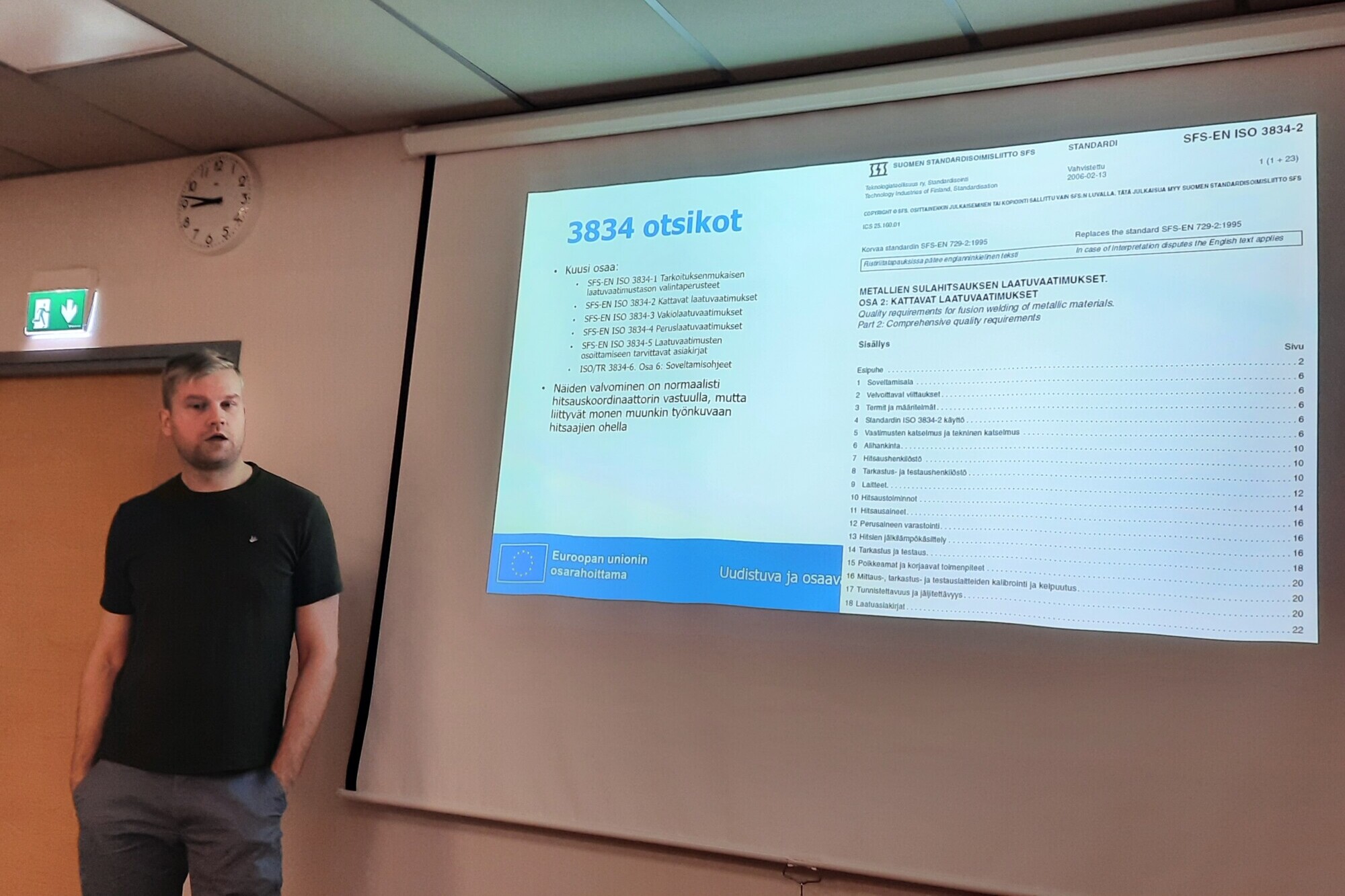 Mies seisoo luokkahuoneessa projisoidun diaesityksen vieressä. Diassa näkyy suomenkielinen teksti 3834 otsikot ja sisällysluettelo, seinällä hätäuloskäyntikyltti.