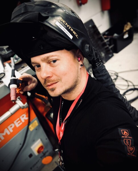 Mustaan hitsaajakypärään ja mustaan paitaan pukeutunut henkilö pitelee hitsauspolttimella oranssin teollisuuslaitteen vieressä työpajassa. Taustalla näkyy kaapeleita ja työkaluja.