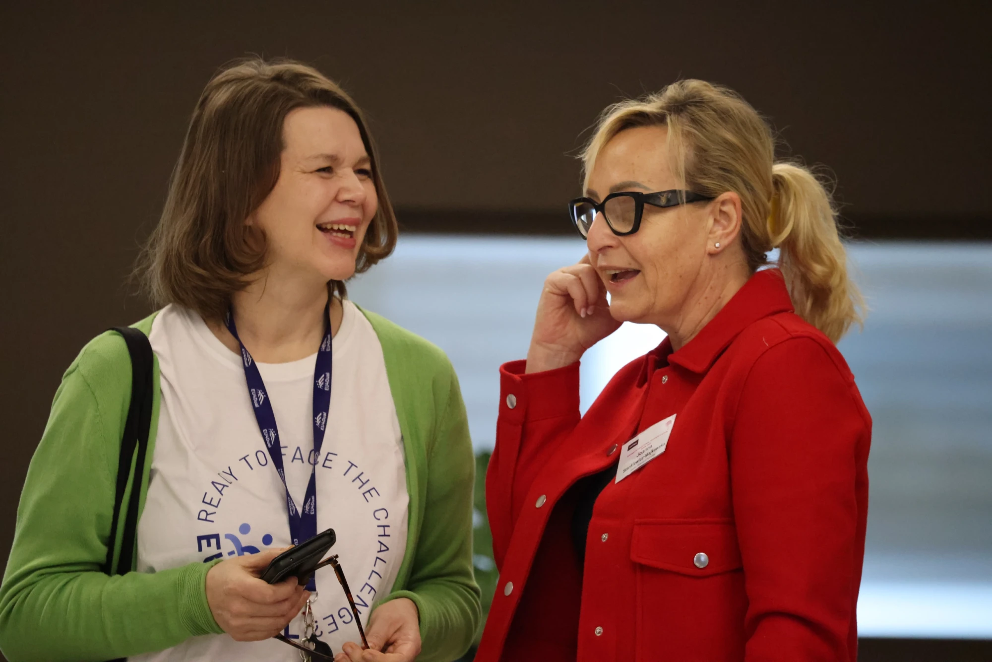 Kaksi naista seisoo ja puhuu; toinen hymyilee, hänellä on vihreä neuletakki ja valkoinen t-paita, kun taas toisella on silmälasit ja punainen takki, jossa on nimikyltti. Molemmat näyttävät käyvän ystävällistä keskustelua sisätiloissa.