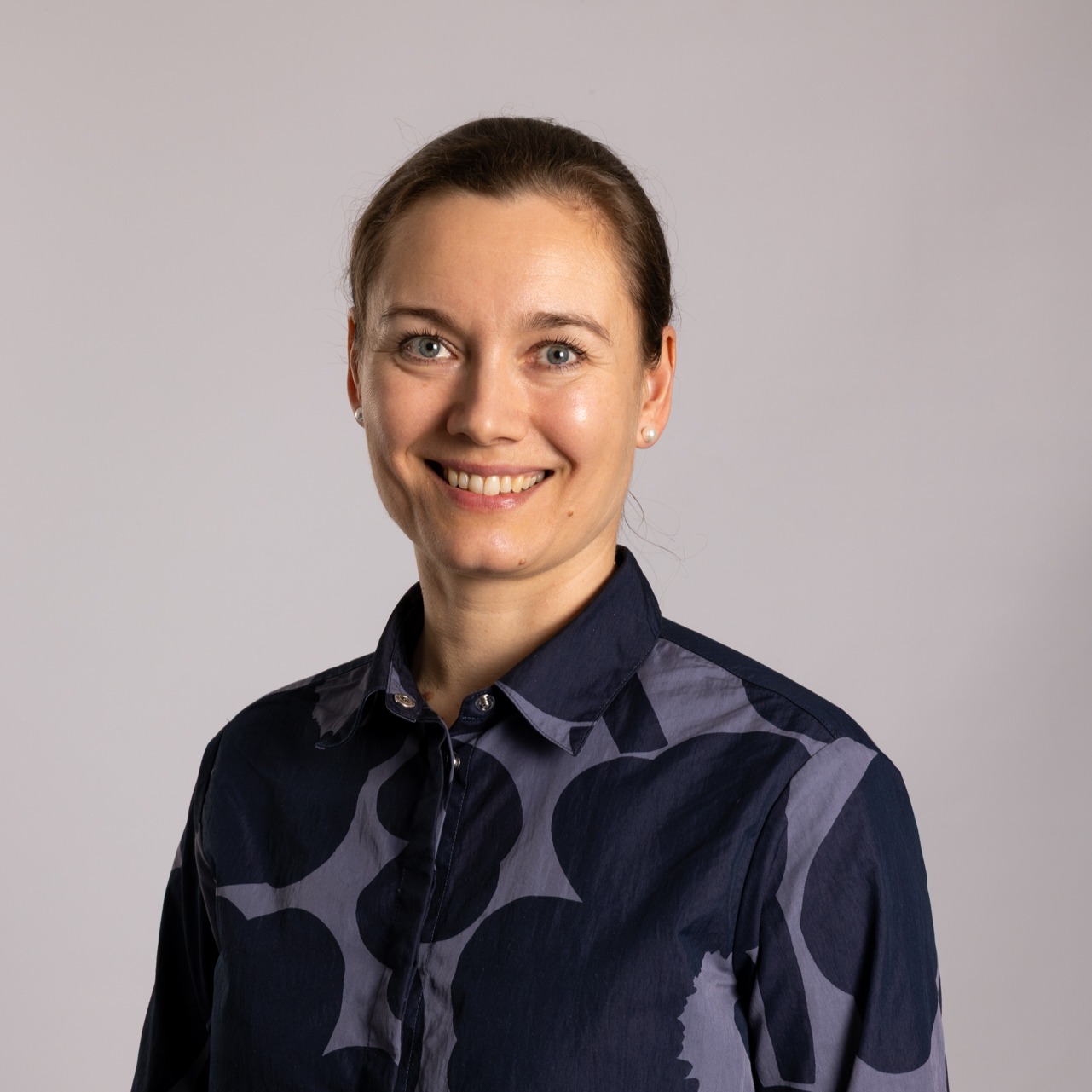Vaaleaihoinen nainen, jolla on taakse sidotut ruskeat hiukset, hymyilee kameralle. Hänellä on yllään tummansininen napitettava paita, jossa on hienovarainen geometrinen kuvio, ja hän seisoo vaaleanharmaata taustaa vasten.