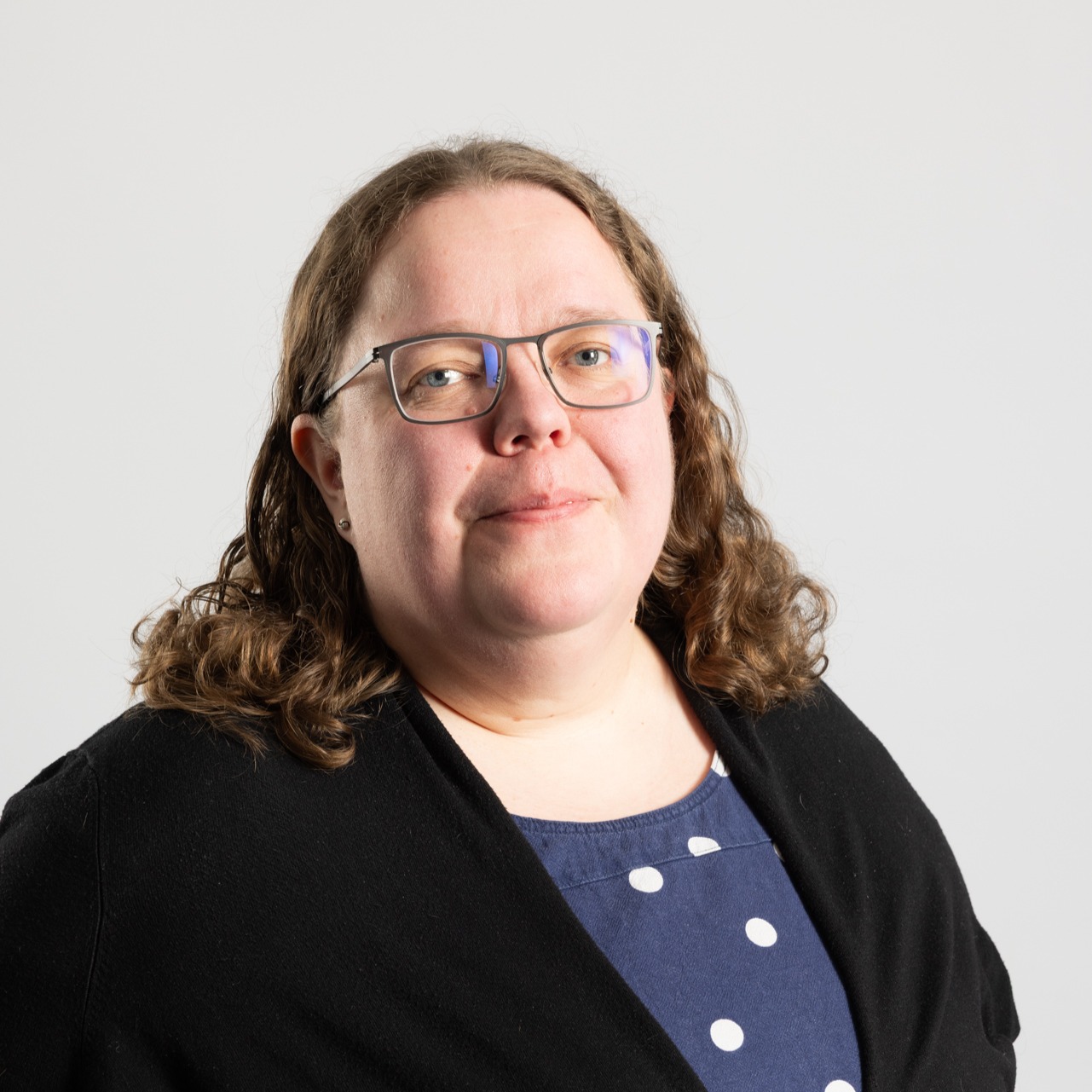 Henkilö, jolla on hartioiden pituiset ruskeat kiharat hiukset, silmälasit, laivastonsininen paita, jossa on valkoisia pilkkuja, ja musta villatakki, poseeraa vaaleanharmaan taustan edessä.