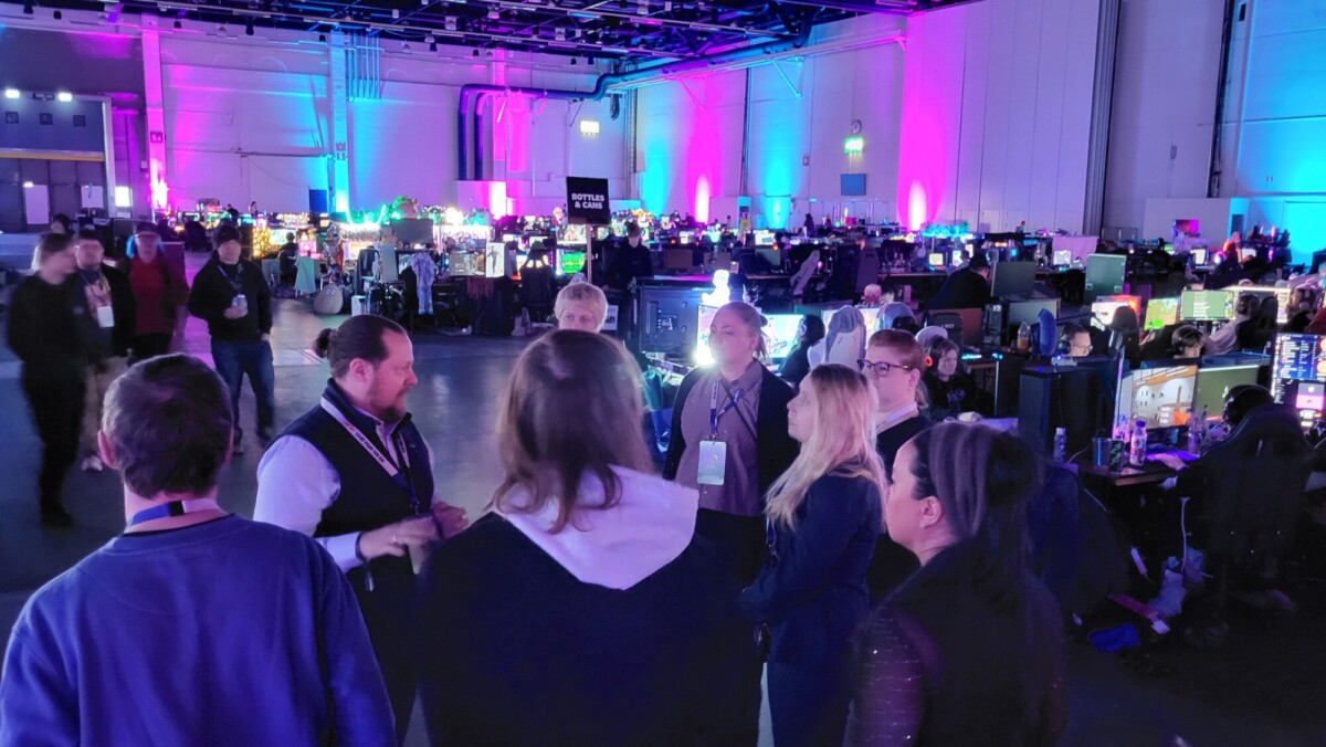 Joukko ihmisiä seisoo piirissä keskustelemassa suuressa sisäpelitapahtumassa, jota ympäröivät tietokonerivit ja värikäs violetti ja sininen valaistus.