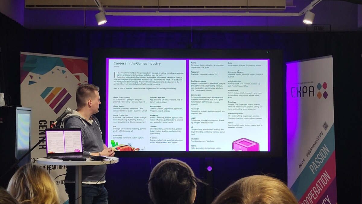 Henkilö seisoo korokkeella pitämässä esitelmää pelialan urasta ja osoittaa tekstiä suurelle näytölle, kun yleisö katsoo. Taustalla näkyy bannereita ja kannettava tietokone.
