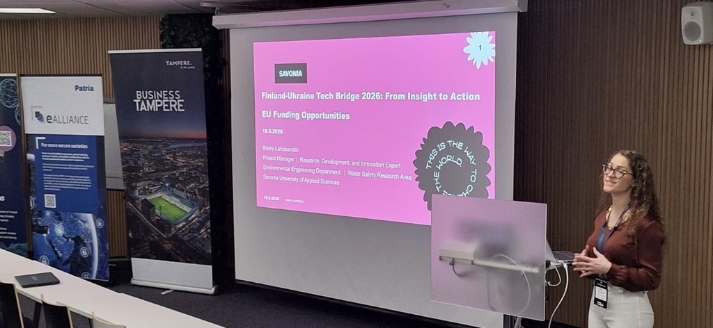 Nainen seisoo korokkeella pitämässä esitelmää Finland-Ukraine Tech Bridge 2024: From Insight to Action EU Funding Opportunities (EU:n rahoitusmahdollisuudet) modernissa konferenssisalissa, jossa on suuri valkokangas ja yleisöistuimet.