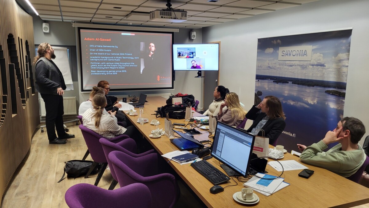 Mies seisoo ja esittelee dioja ryhmälle, joka istuu neuvottelupöydän ympärillä kannettavien tietokoneiden, muistilehtiöiden ja kahvikuppien kanssa. Näytöllä näkyy videopuhelu, ja toinen projektio näyttää esityksen yksityiskohdat seinällä.