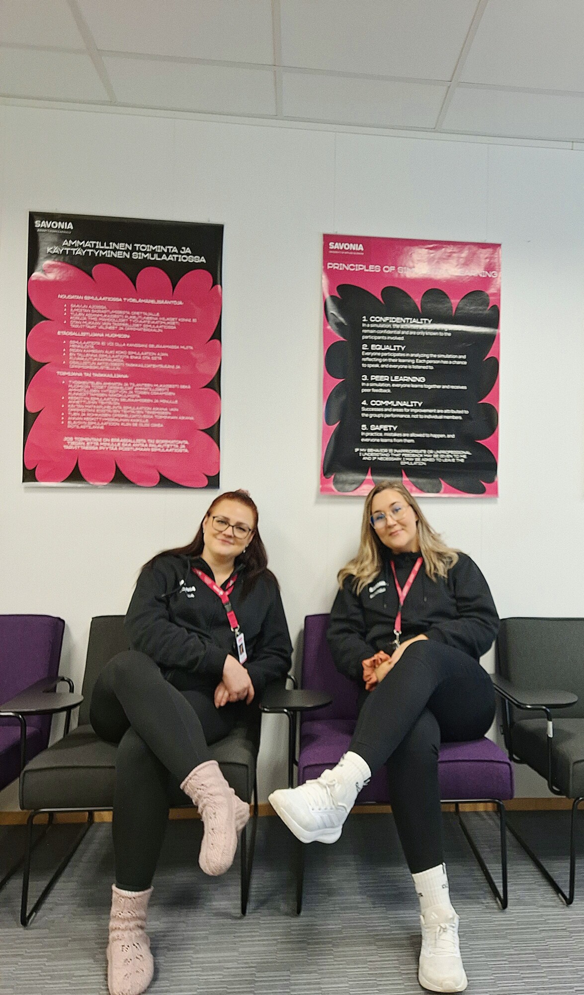 Kaksi naista, joilla on mustat takit, vaaleanpunaiset nauhat ja silmälasit, istuu vierekkäin tuoleilla odotustilassa. Heidän takanaan on kaksi vaaleanpunaisia ja mustia kuvioita sisältävää julistetta valkoisella seinällä.