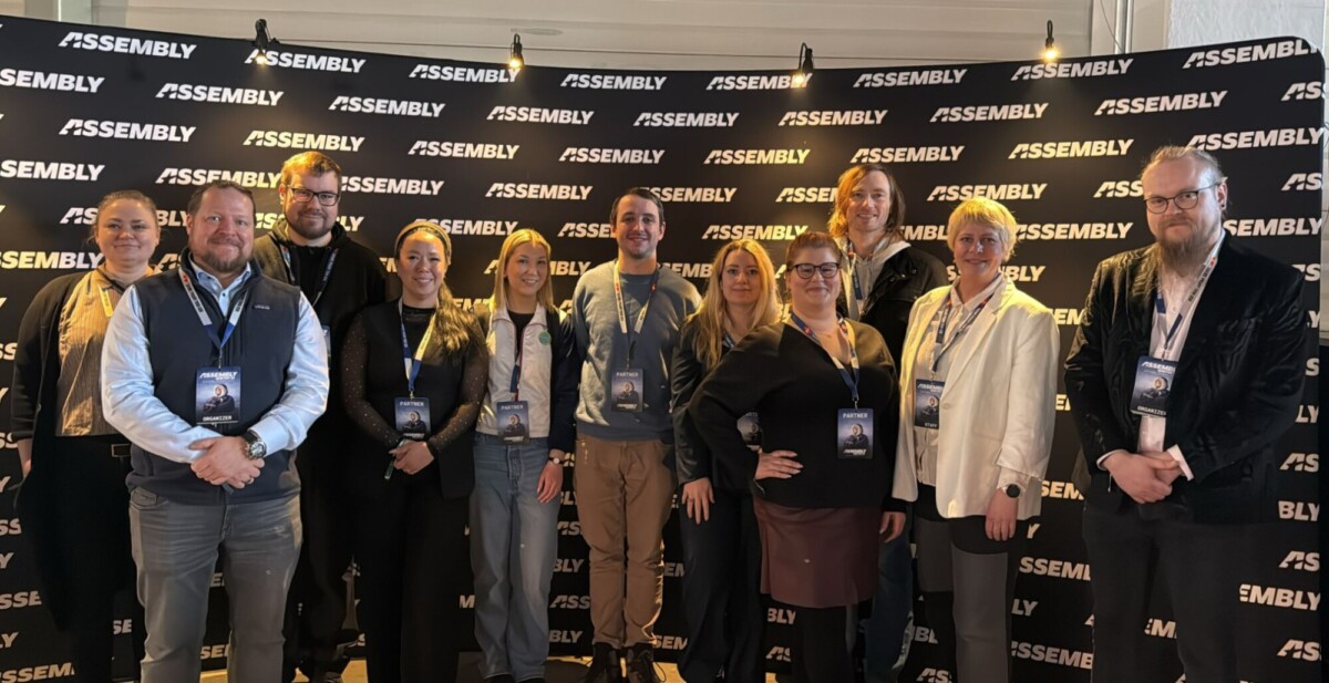 Kaksitoista hymyilevää ihmistä seisoo yhdessä mustan taustan edessä, jossa on toistuvia valkoisia ASSEMBLY-logoja. Useimmilla on konferenssimerkit ja liikemiesten vaatteet.