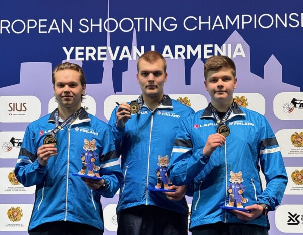 Kolme sinisiin Suomi-takkeihin pukeutunutta miesurheilijaa seisoo palkintokorokkeella mitalit ja pokaalit kädessään ampumaurheilun Euroopan mestaruuskilpailuissa Jerevanissa, Armeniassa, ja taustalla on tapahtuman ja paikan tiedot.
