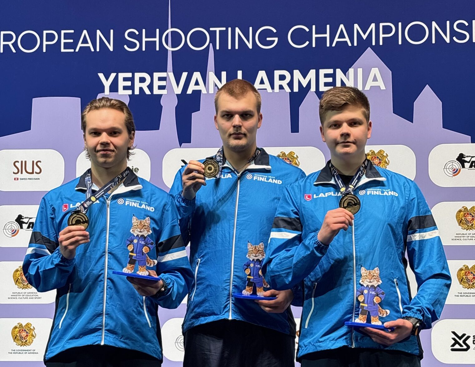 Kolme sinisiin Suomi-takkeihin pukeutunutta miesurheilijaa seisoo palkintokorokkeella mitalit ja pokaalit kädessään ampumaurheilun Euroopan mestaruuskilpailuissa Jerevanissa, Armeniassa, ja taustalla on tapahtuman ja paikan tiedot.