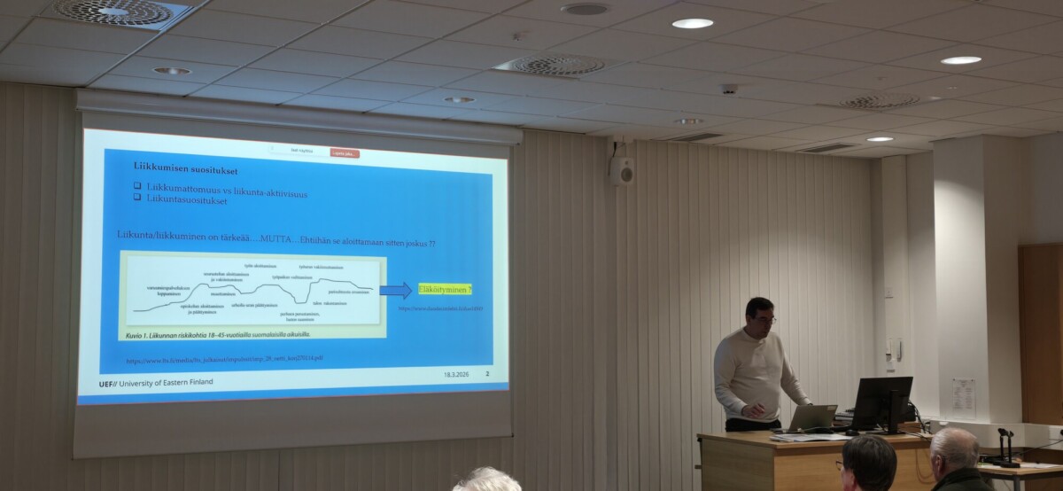 Mies seisoo korokkeella pitämässä esitelmää yleisölle. Hänen takanaan olevan modernin luentosalin seinälle heijastetaan sinisellä pohjalla oleva dia, suomenkielinen teksti ja kaavio.