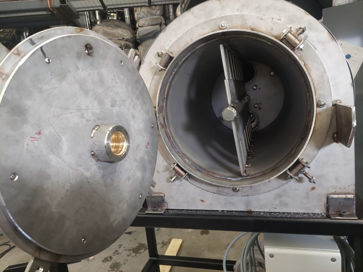 Suuri, lieriömäinen, metallinen teollisuuskone, jonka pyöreä ovi on auki ja josta paljastuu sisätila, jossa on metallisia teriä ja lamelleja. Ulkopinnalla on jonkin verran kulumaa, ja kone on kiinnitetty mustaan kehykseen.