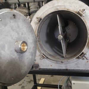 Suuri, lieriömäinen, metallinen teollisuuskone, jonka pyöreä ovi on auki ja josta paljastuu sisätila, jossa on metallisia teriä ja lamelleja. Ulkopinnalla on jonkin verran kulumaa, ja kone on kiinnitetty mustaan kehykseen.