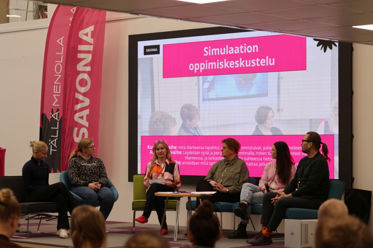 Viisi ihmistä istuu lavalla yleisön edessä paneelikeskustelussa. Heidän takanaan on suuri valkokangas, jolla näkyy suomenkielinen teksti Simulaation oppimiskeskustelu ja valokuvia.