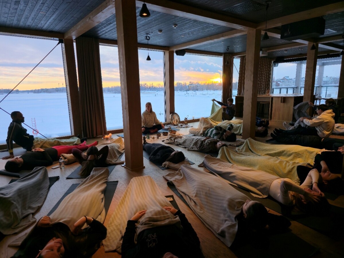 Ryhmä ihmisiä makaa lattialla peittojen alla viihtyisässä huoneessa, jossa on suuret ikkunat auringonlaskun aikaan. Ohjaaja istuu edessä rummun kanssa ja johtaa rentouttavaa tai meditatiivista istuntoa. Ulkona näkyy luminen maisema.