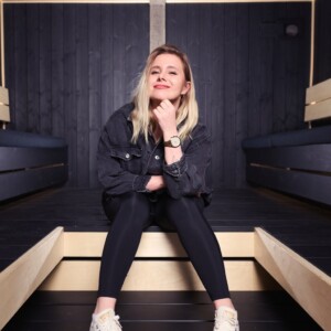 Vaaleatukkainen nainen, jolla on musta takki, mustat legginsit ja valkoiset lenkkarit, istuu puisella korokkeella modernissa, tummaksi paneloidussa huoneessa hymyillen kevyesti ja nojaten leuka kädelleen.