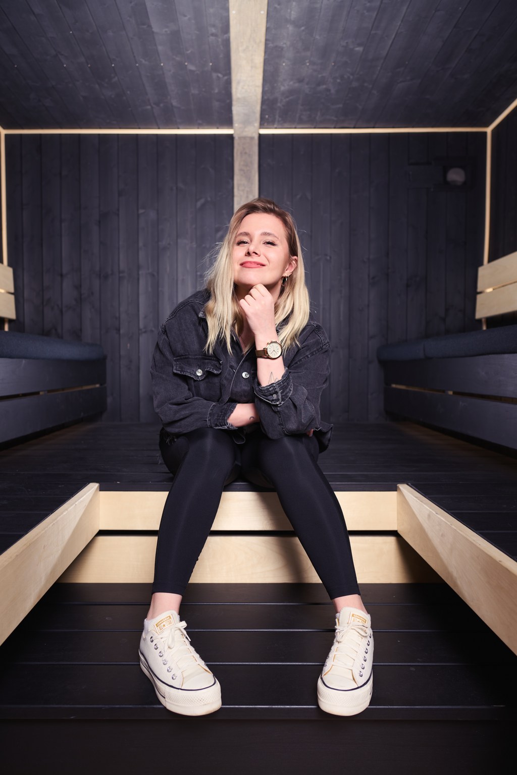 Vaaleatukkainen nainen, jolla on musta takki, mustat legginsit ja valkoiset lenkkarit, istuu puisella korokkeella modernissa, tummaksi paneloidussa huoneessa hymyillen kevyesti ja nojaten leuka kädelleen.