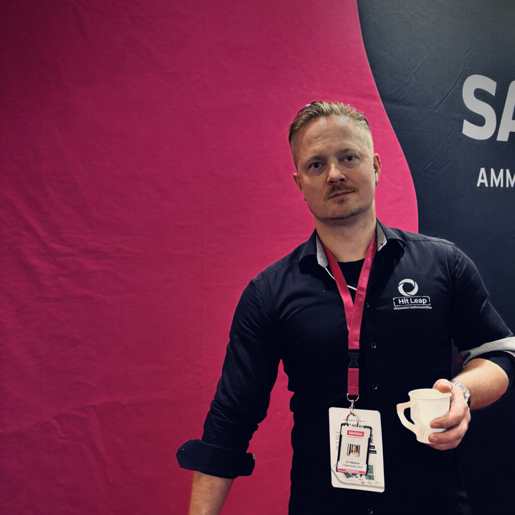 Vaaleahiuksinen mies, jolla on musta paita ja konferenssimerkki, seisoo vaaleanpunaisen ja mustan taustan edessä, pitää valkoista kuppia kädessään ja katsoo kameraan.