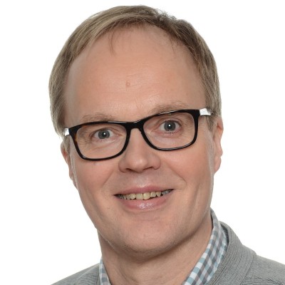 Mies, jolla on lyhyet vaaleanruskeat hiukset, mustat suorakulmaiset silmälasit, harmaa pusero ja ruutupaita, hymyilee kameralle valkoista taustaa vasten.