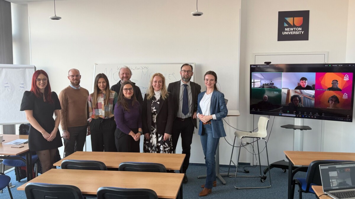 Kahdeksan hengen ryhmä seisoo ja hymyilee luokkahuoneessa Newtonin yliopistossa, ja neljä muuta osallistujaa näkyy videopuhelussa oikealla olevalla näytöllä. Luokkahuoneen kalusteet ja valkotaulu ovat näkyvissä.