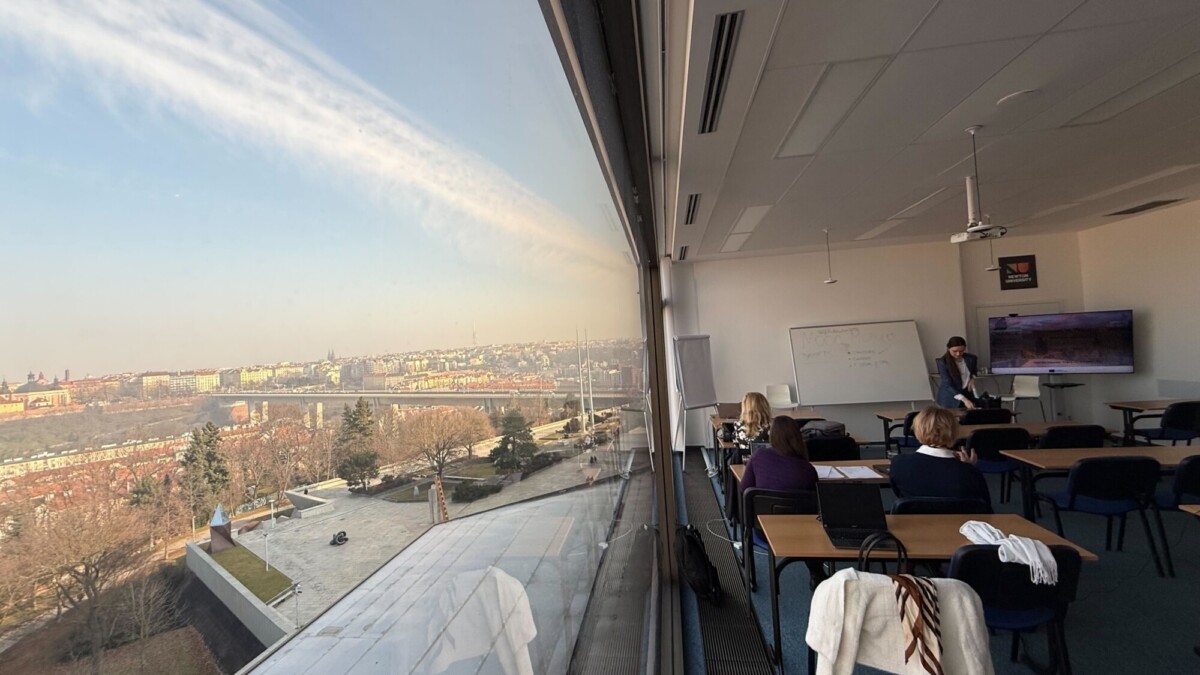 Suuri luokkahuone, jossa ihmiset istuvat pöydissä ja juontaja on taulun ääressä. Korkea ikkuna heijastaa sekä oppituntia että panoraamanäkymää kaupunkiin, jossa on rakennuksia, puita ja joki ulkona.