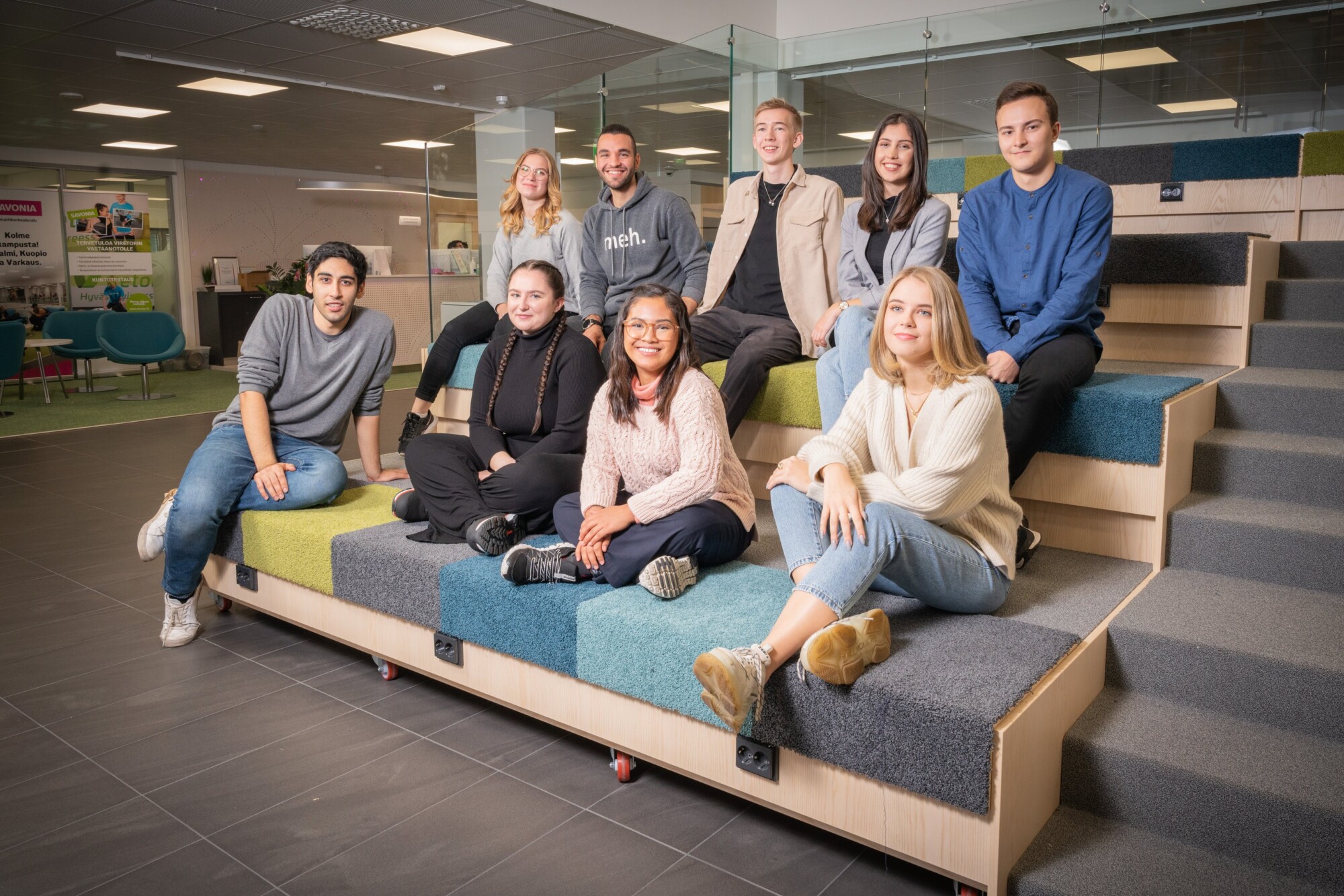 Yhdeksän nuoren aikuisen ryhmä istuu ja hymyilee modernilla porrastetulla istuimella valoisassa, avoimessa sisätilassa, jonka taustalla näkyvät lasiseinät ja toimistokalusteet.