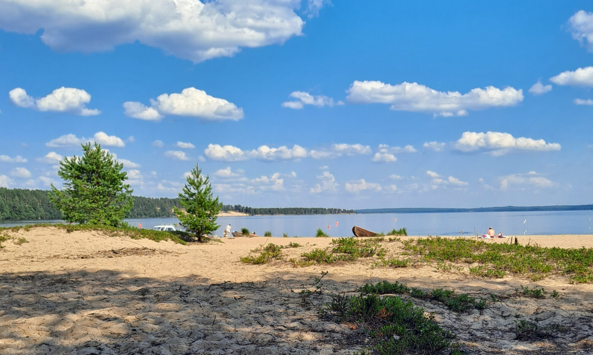 Hiekkaranta, jossa on vihreitä pensaita ja pieniä mäntyjä ja josta avautuu näkymä rauhalliselle järvelle kirkkaansinisen taivaan alla, jossa on hajanaisia valkoisia pilviä. Muutama ihminen rentoutuu veden äärellä nauttien aurinkoisesta päivästä.