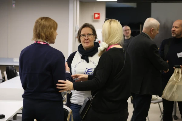 Kolme naista seisoo neuvotteluhuoneessa keskustelemassa, ja yhdellä heistä on valkoinen t-paita ja silmälasit. Muut ihmiset keskustelevat taustalla. Kaikki näyttävät olevan mukana keskustelussa.