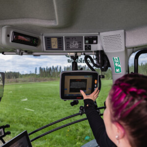 Vaaleanpunaiset hiukset omaava henkilö koskettaa modernin traktorin ohjaamossa kosketusnäyttöä, ja tuulilasin läpi näkyy vihreitä peltoja ja puita. Kojelaudan yläpuolelle on asennettu erilaisia ohjauspaneeleita.