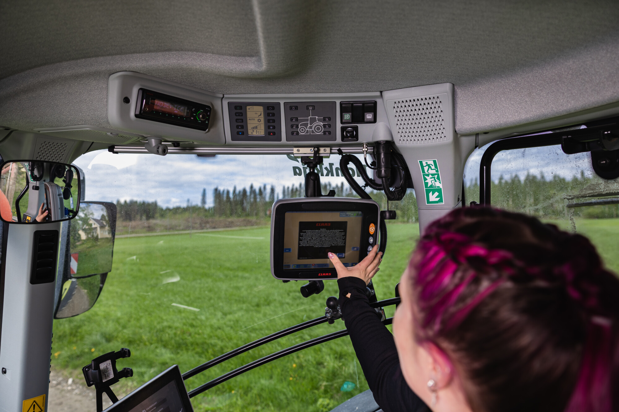 Vaaleanpunaiset hiukset omaava henkilö koskettaa modernin traktorin ohjaamossa kosketusnäyttöä, ja tuulilasin läpi näkyy vihreitä peltoja ja puita. Kojelaudan yläpuolelle on asennettu erilaisia ohjauspaneeleita.