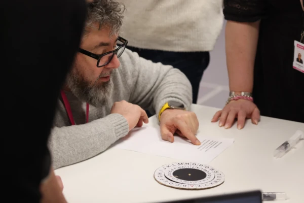 Silmälasipäinen parrakas henkilö osoittaa pöydällä olevaa paperia, jota muut ympäröivät. Pöydällä on myös salakirjoituspyörä, jossa on kirjaimia ympyrän sisällä. Keskitytään ilmeisesti arvoituksen tai koodin ratkaisemiseen.