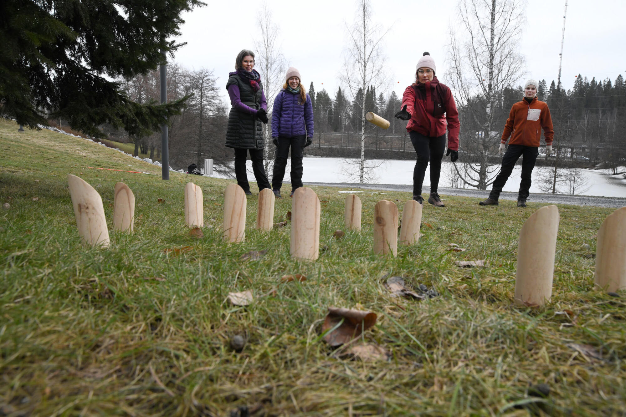 Neljä ihmistä pelaa ulkona puisilla nastoilla ja kapulalla nurmikolla järven lähellä puistossa, taustalla puita ja pilvinen taivas. Yksi henkilö on kesken heiton ja tähtää keiloja.