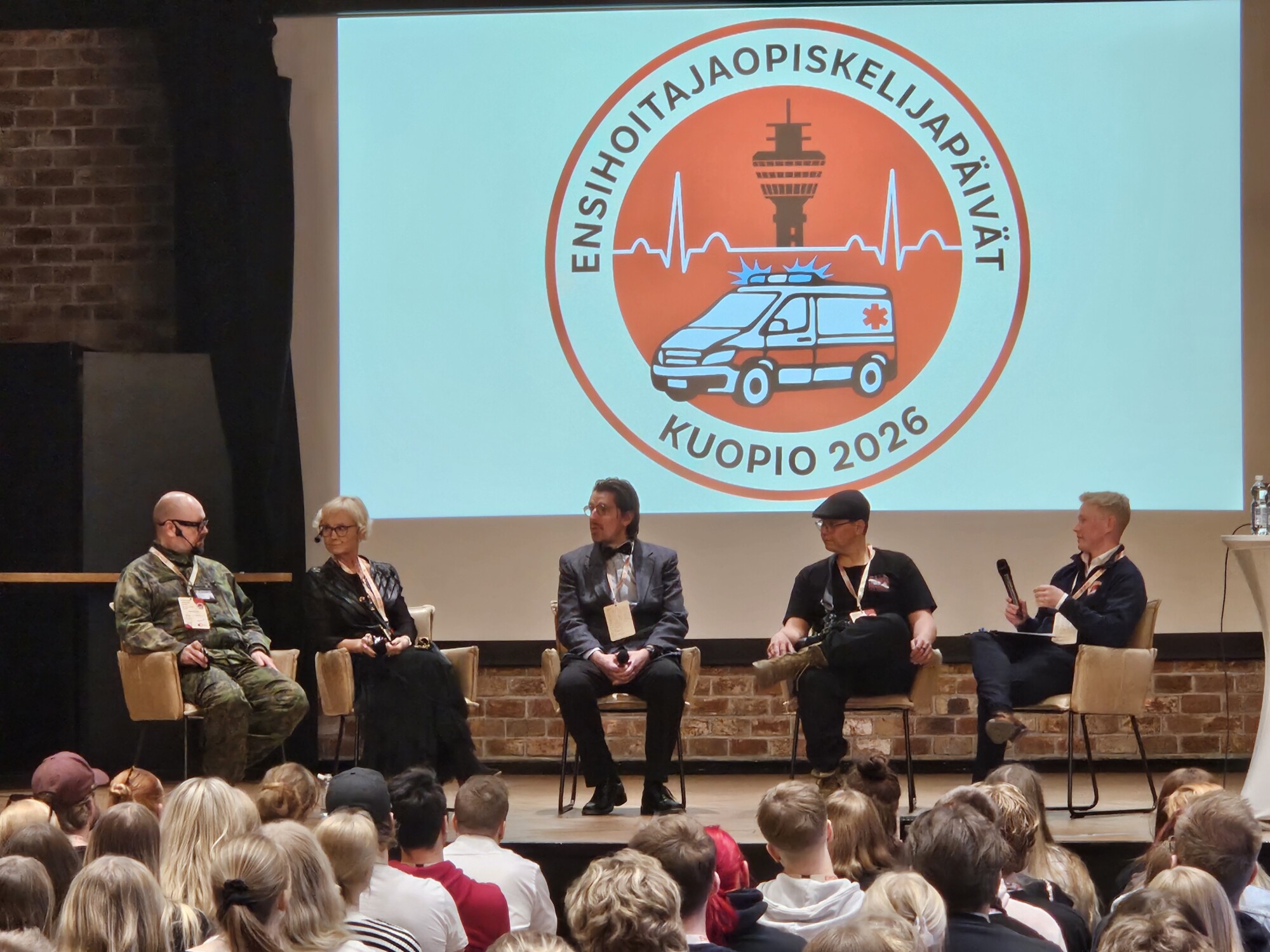 Viisi ihmistä istuu lavalla yleisön edessä paneelikeskustelussa. Heidän takanaan olevassa suuressa logossa lukee Ensihoitajaopiskelijapäivät Kuopio 2026 ja siinä on ambulanssigrafiikka.