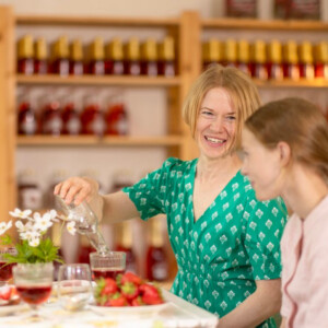 Kaksi naista istuu pöydässä, jossa on mansikoita, juomia ja kukkia. Toinen vihreäpukuinen nainen kaataa vettä hymyillen. Taustalla on hyllyjä, joissa on pulloja. Logot näkyvät vasemmassa yläkulmassa.